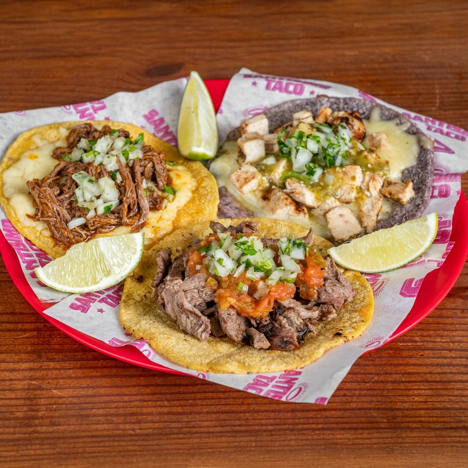 Three Mexican tacos topped with shredded beef, grilled chicken, onions, cilantro, salsa, melted cheese, and lime wedges on a red plate.