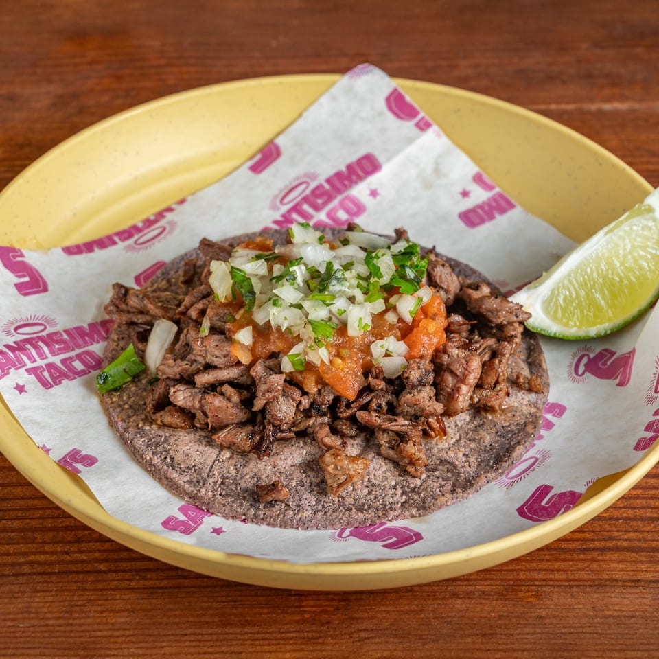 A blue-corn tortilla topped with grilled meat, onions, cilantro, salsa, and a lime wedge on a yellow plate.