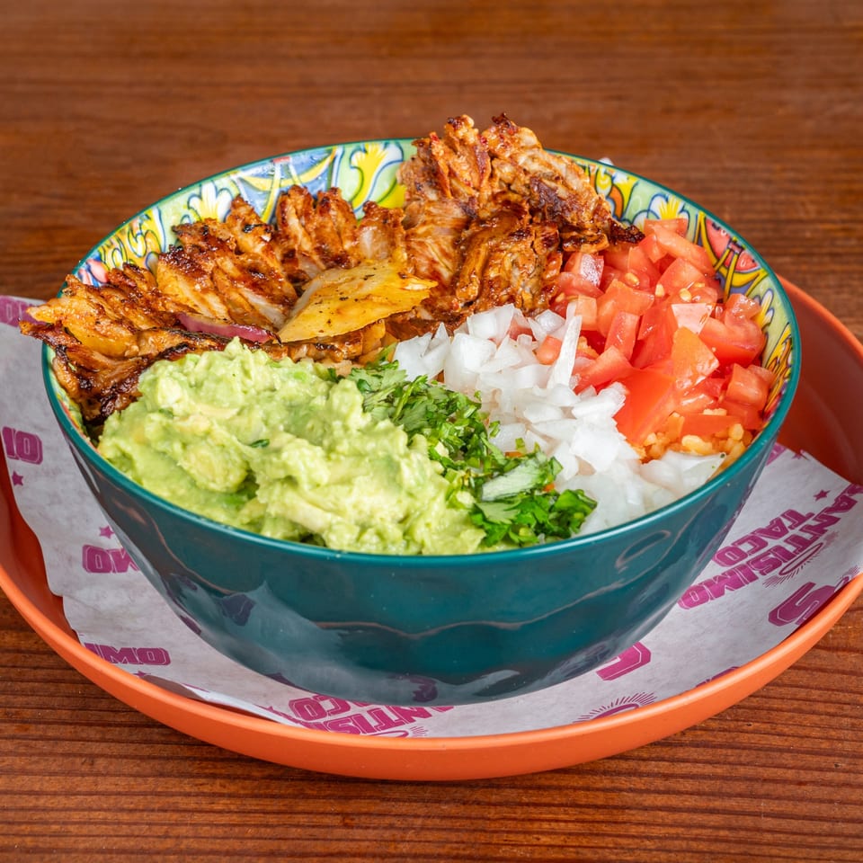 Colorful bowl of grilled meat topped with guacamole, diced tomatoes, onions, cilantro, and roasted pineapple on an orange tray.