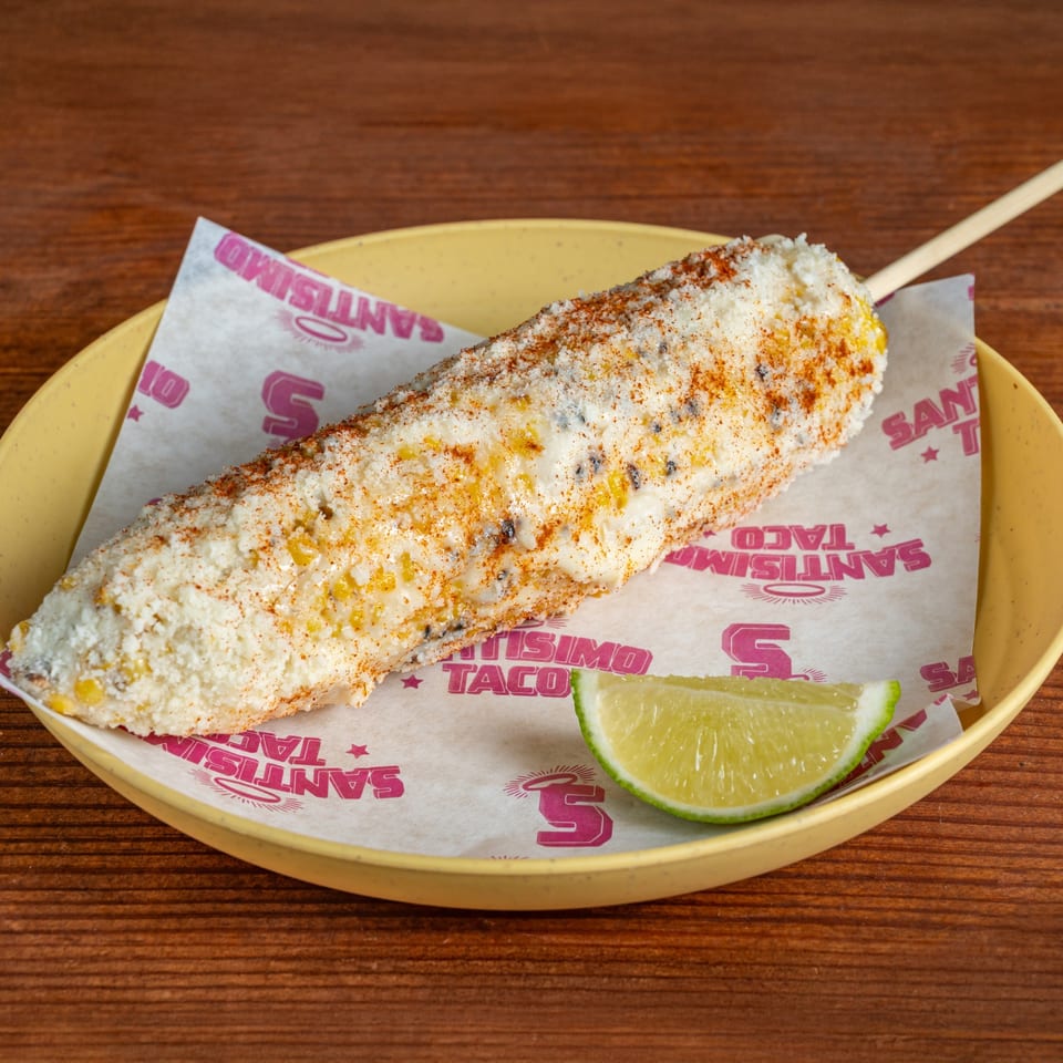 Mexican street corn coated with cheese and chili seasoning, served on a branded plate with a lime wedge.