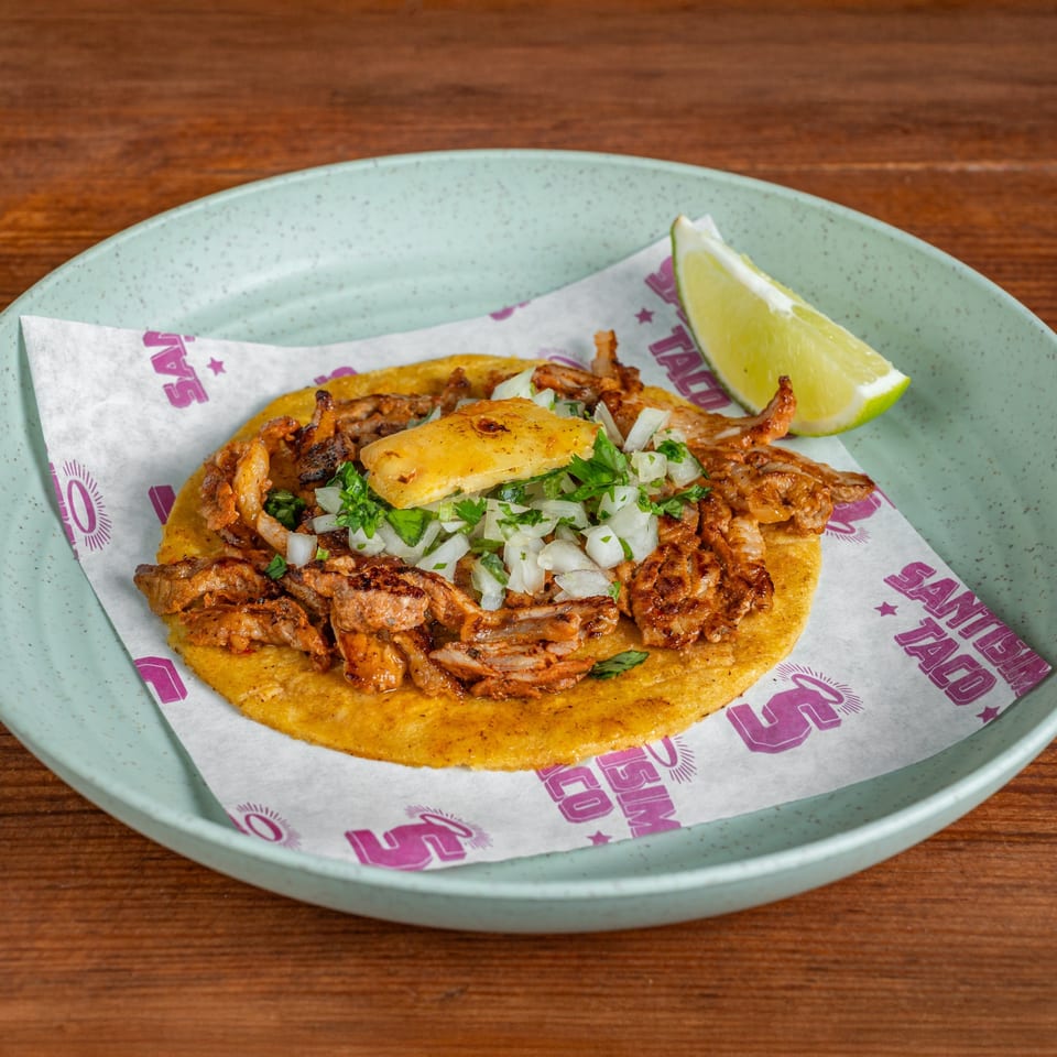 Mexican-style corn tortilla topped with grilled meat, chopped onion, cilantro, pineapple, and a lime wedge on a plate.